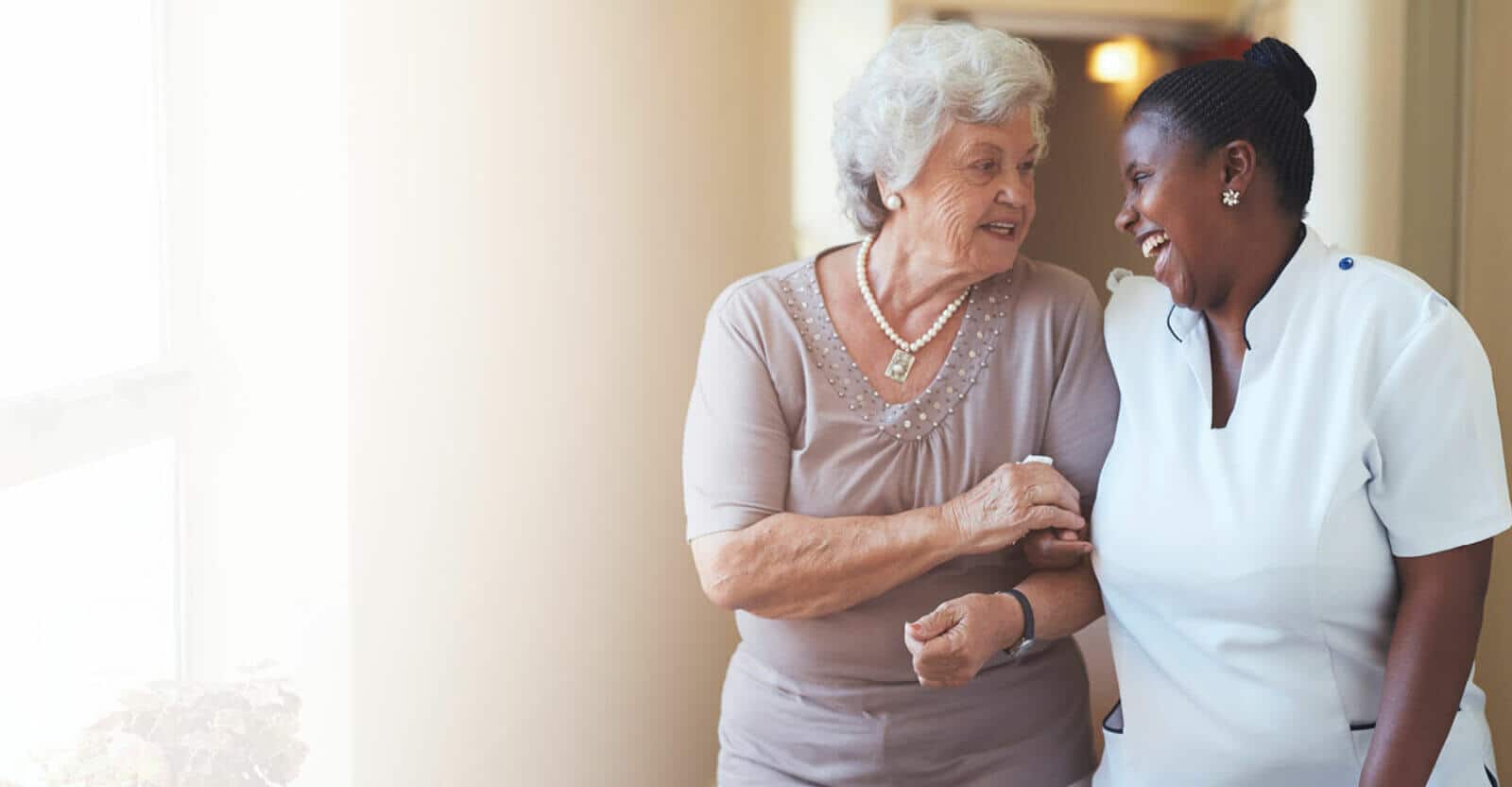 Older woman walking down the hall with a nurse, laughing and holding hands